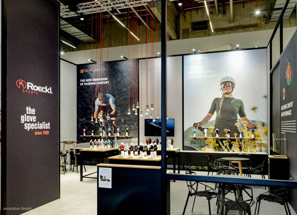 Roeckl Messestand mit beleuchteten Fahrradexponaten, dunklem Standdesign und großformatigen Sportler-Grafiken